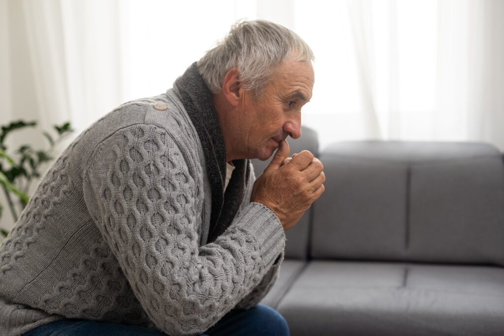 Ein älterer Mann in einem grauen Pullover sitzt nach vorne gebeugt auf einem Sofa, hat die Hände vor dem Mund gefaltet und blickt nachdenklich.