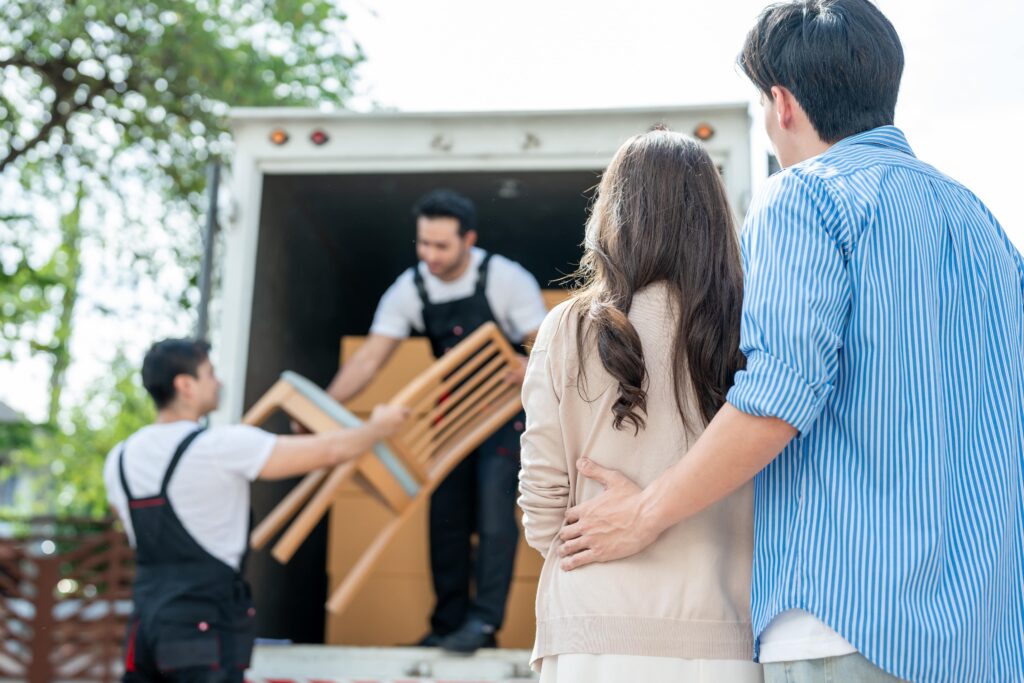 
Ein Paar beobachtet an einem sonnigen Tag, wie zwei Möbelpacker die Holzmöbel in einen Lastwagen laden.