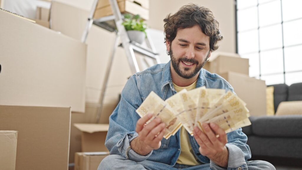 Ein Mann, der auf dem Boden zwischen Pappkartons sitzt, hält mehrere 100-Euro-Scheine in der Hand und fächert sie auf, während er das Geld lächelnd betrachtet. Er konnte anscheinend seine Umzugskosten steuerlich absetzen.