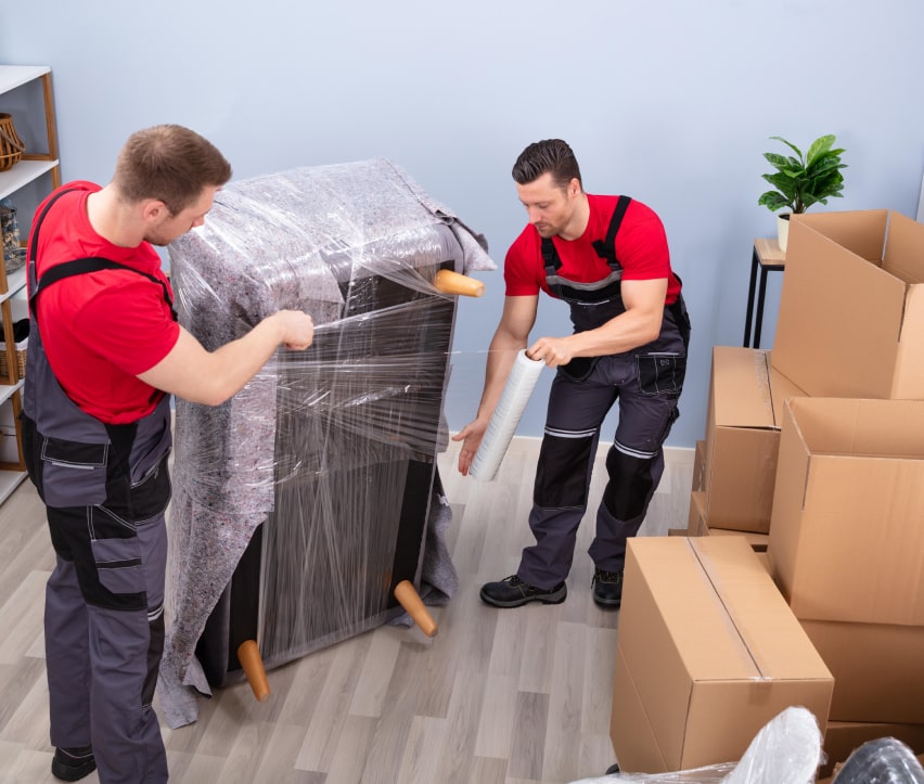 Zwei Umzugshelfer in roten Hemden und Latzhosen wickeln in einem mit Kartons und einigen Zimmerpflanzen gefüllten Raum Möbel in Plastikfolie ein.
