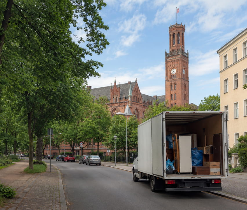 In Berlin-Lichtenberg steht ein Umzugswagen mit geöffneten Hecktüren in einer ruhigen, von Bäumen und Gebäuden gesäumten Straße; im Hintergrund ist ein großer Kirchturm zu sehen.