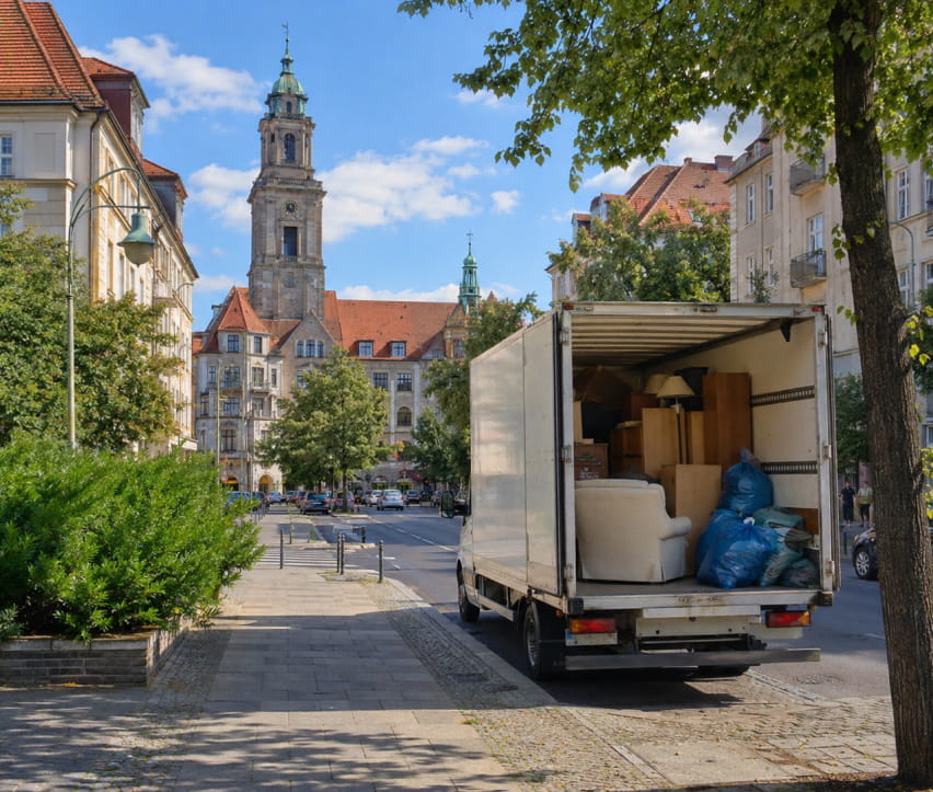 Ein Umzugswagen mit offenen Türen , bereit für eine Entrümpelung in Steglitz, steht auf einer Schloßstraße, beladen mit Kisten und Möbeln und blauen Säcken, im Hintergrund ist das Schloß Steglitz zu sehen.