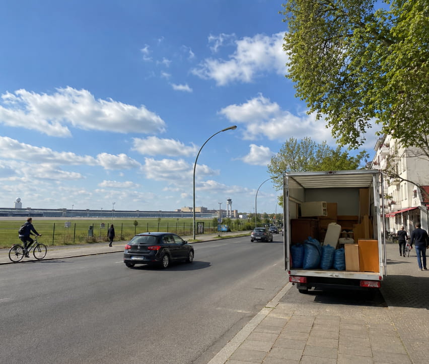 Ein Umzugswagen mit Möbeln und Koffern für eine Entrümpelung steht auf einer Straße in der Nähe des Tempelhofer Felds; unter einem teilweise bewölkten Himmel sind Autos, ein Radfahrer und Fußgänger zu sehen.