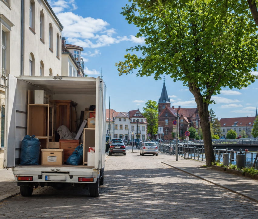 Ein Umzugswagen mit offenen Türen und beladenen Kisten steht an einem sonnigen Tag auf einer von Bäumen und Gebäuden gesäumten Kopfsteinpflasterstraße für eine Entrümpelung in Berlin-Treptow-Köpenick, in der Nähe des Ufers.