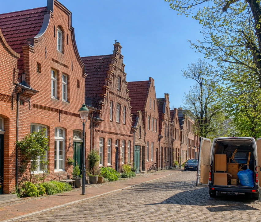 Eine Reihe von Backsteinhäusern mit Satteldächern säumt eine Kopfsteinpflasterstraße im hollaendischen Viertel in Potsdam; dort steht ein weißer Transporter mit geöffneten Hecktüren, der teilweise mit Kisten und Möbeln wegen einer Entrümpelung beladen ist.
